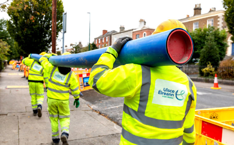 Oibritheoirí an chláir laghdaithe sceite agus píobán á iompar acu ar feadh bóthair